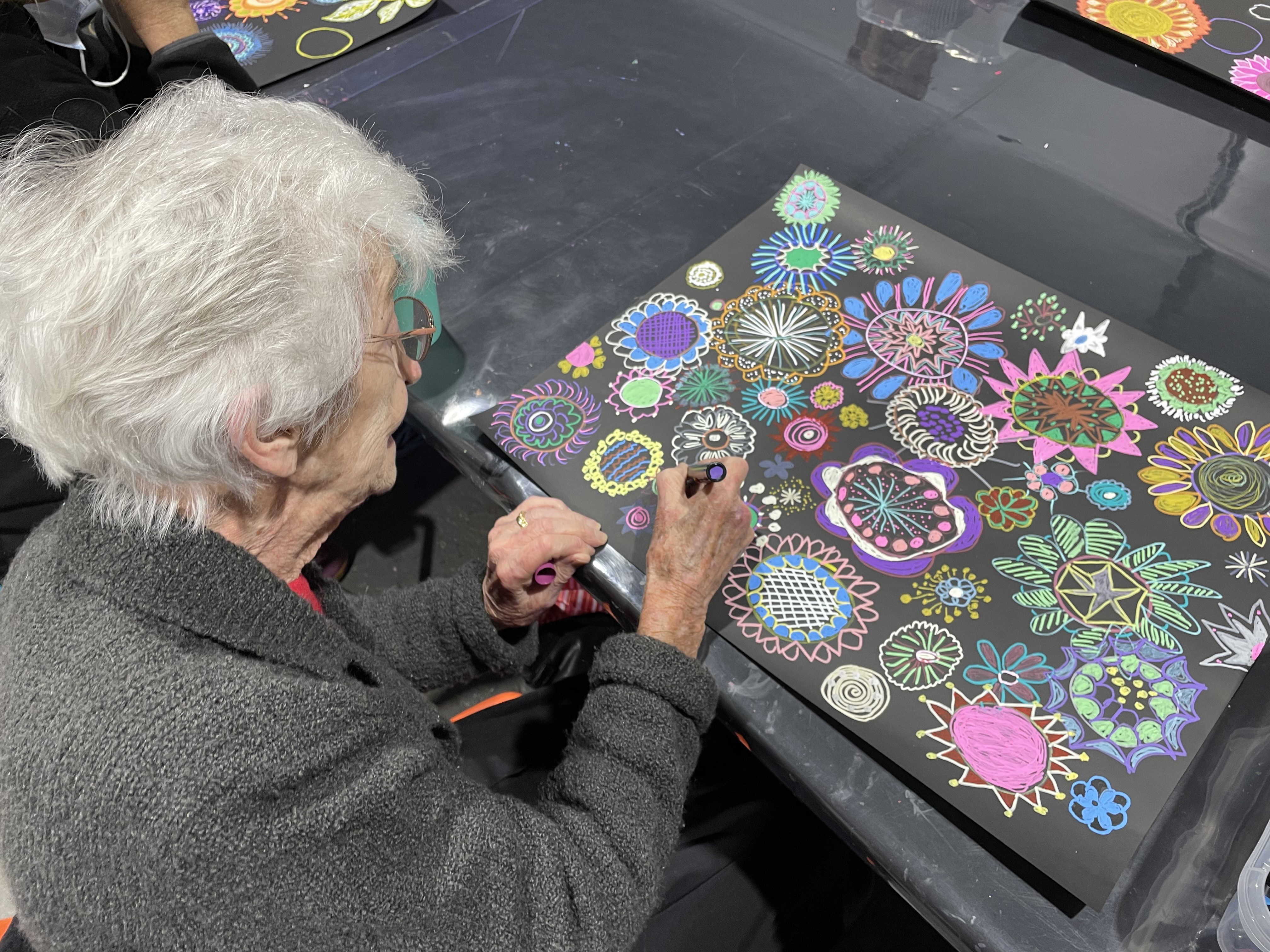 老妇人拿着笔，在一张大黑纸上画出鲜艳的五颜六色的花朵。